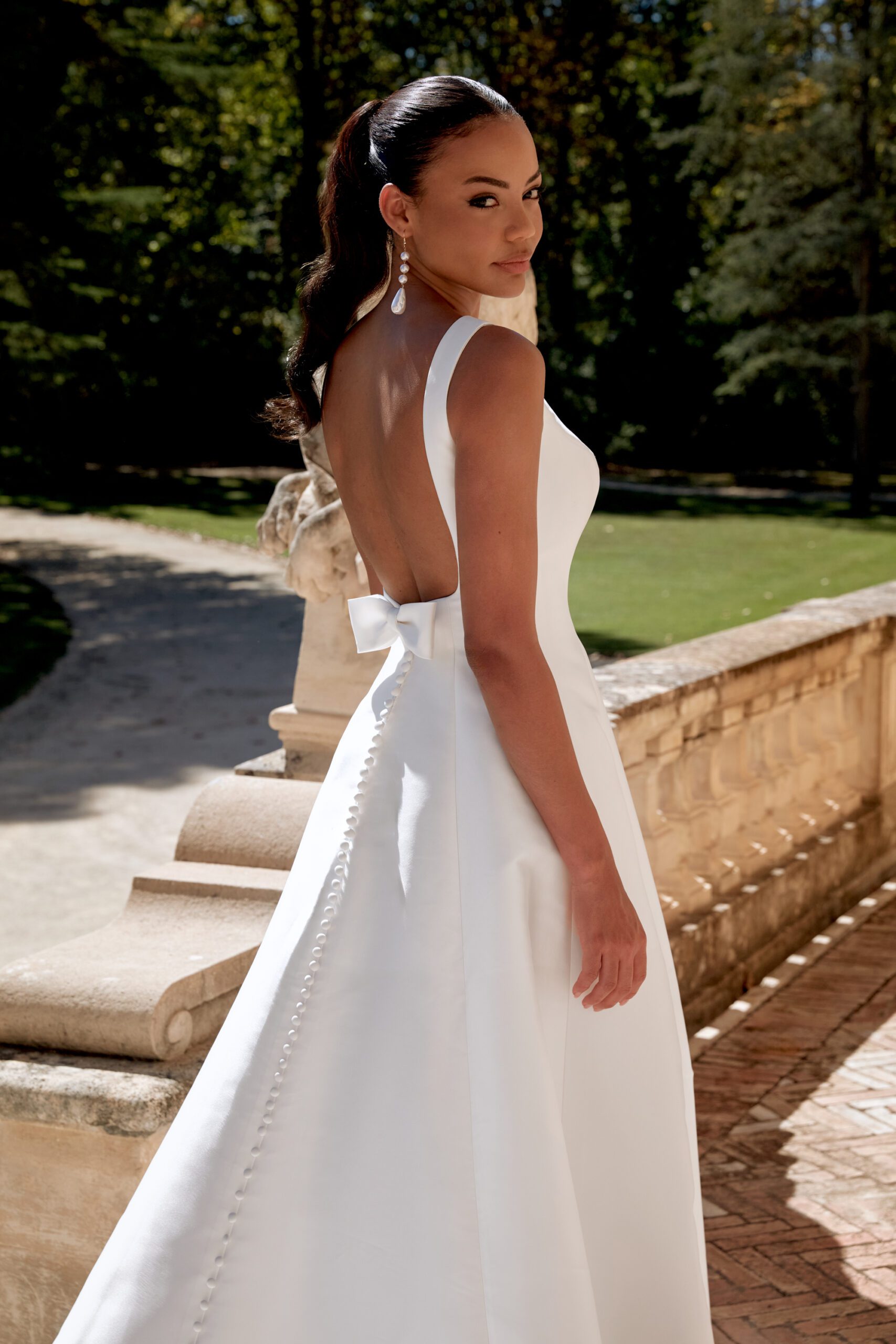 Das Brautkleid ist aus Satin und hat einen U-Boot Ausschnitt. Der tiefe Rückenausschnitt wird mit einer Schleife und einer Knopfleiste abgerundet.